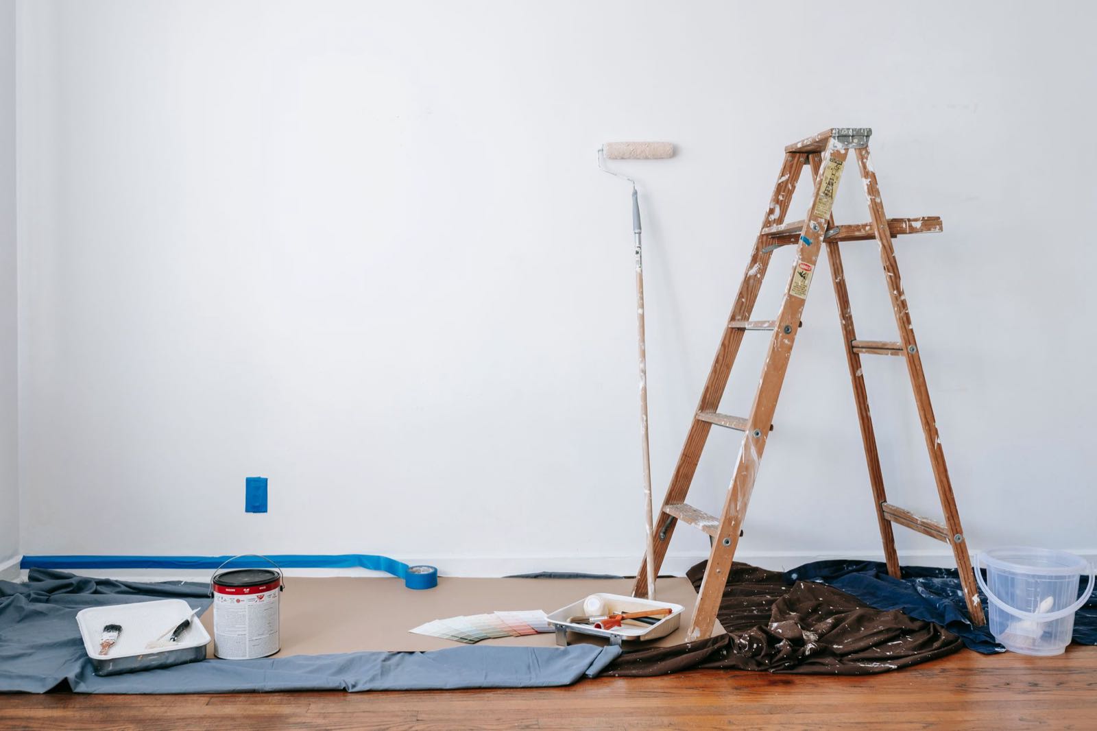 Empty room prepared for interior painting, with taped baseboards, drop cloths, a wooden ladder, and a paint roller